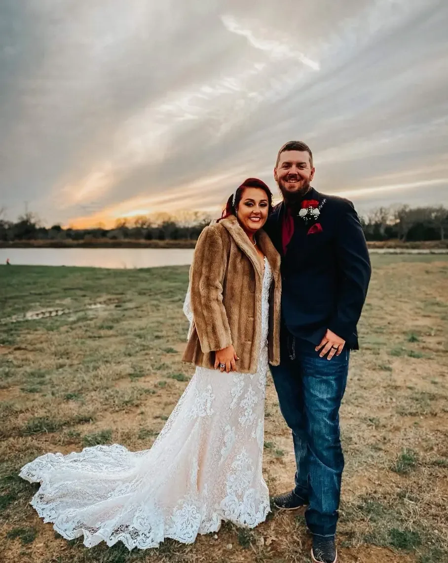 Bride in a white fur wrap and groom in a dark suit posing at sunset beside the lake at Reverie at Diamond D in Normangee, Texas