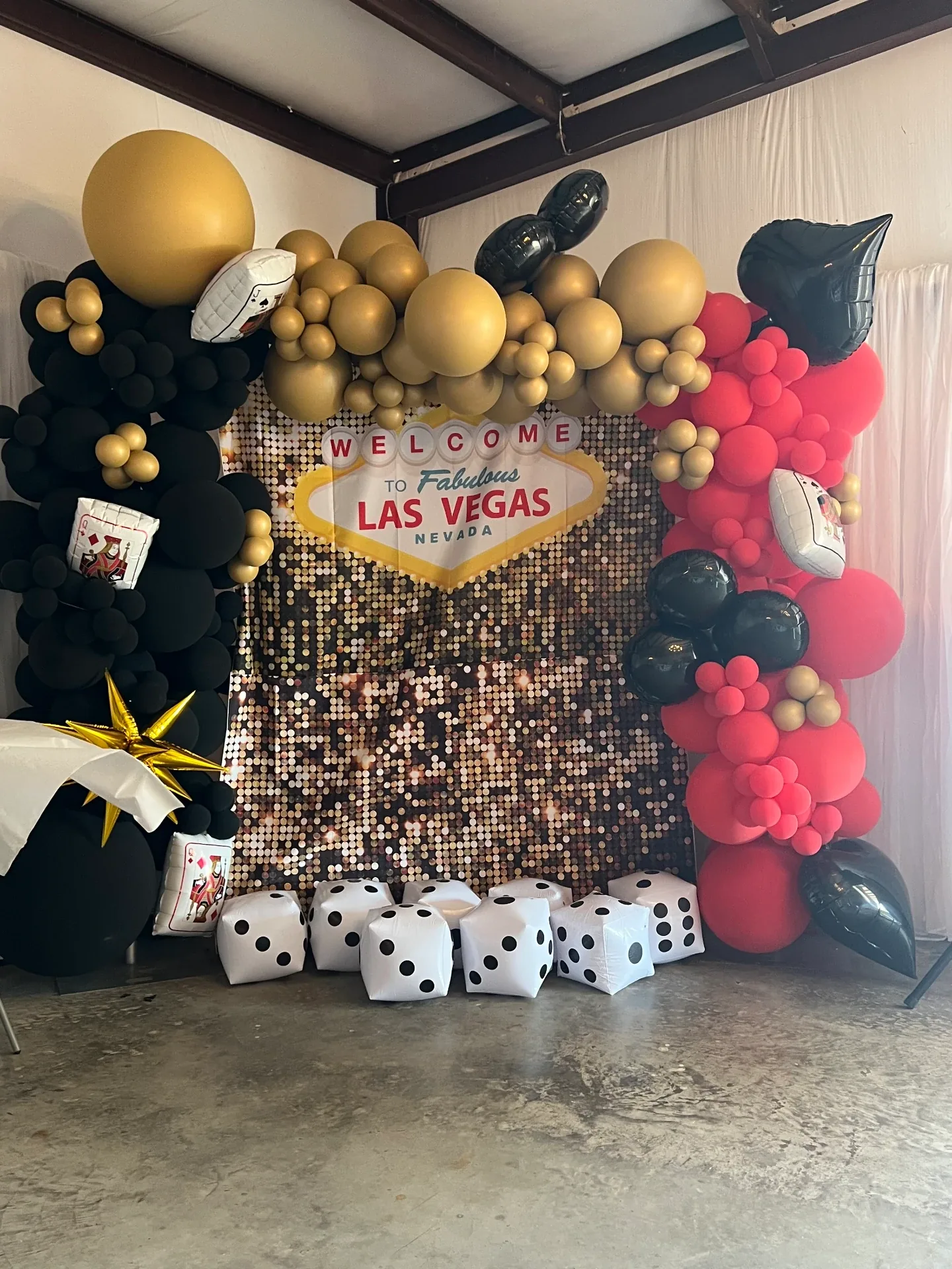 Gold sequin photo backdrop with a Welcome to Las Vegas sign, black and red balloon garland and inflatable dice at the Love Like Bailin event