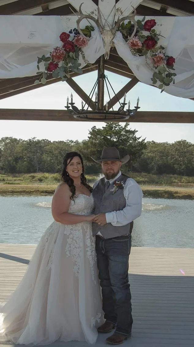 Bride and groom standing beneath a floral and antler arch under the lakeside pavilion at Reverie at Diamond D