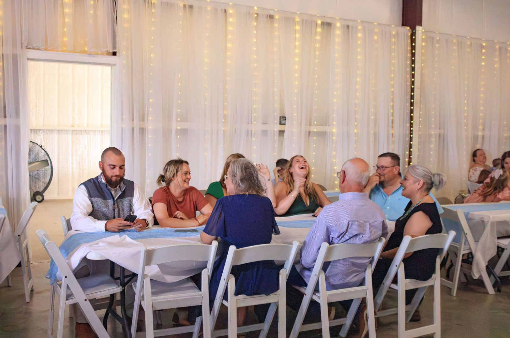 Jaelyn & Keelan's wedding at Reverie at Diamond D — guests laughing at a reception table beneath the string lights