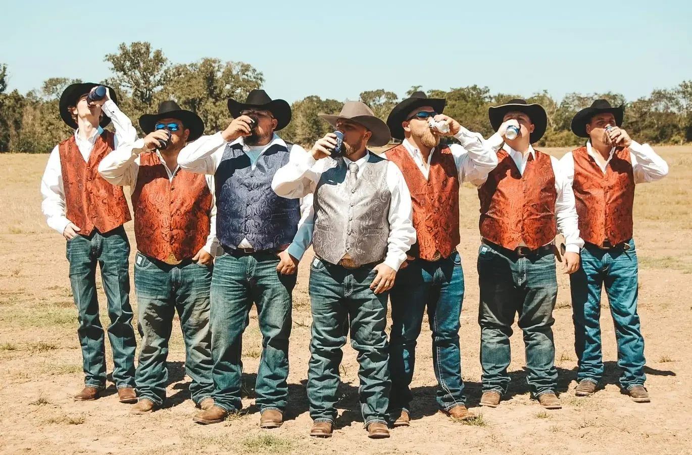 Seven groomsmen in cowboy hats and orange paisley vests raising a synchronized toast in a ranch field