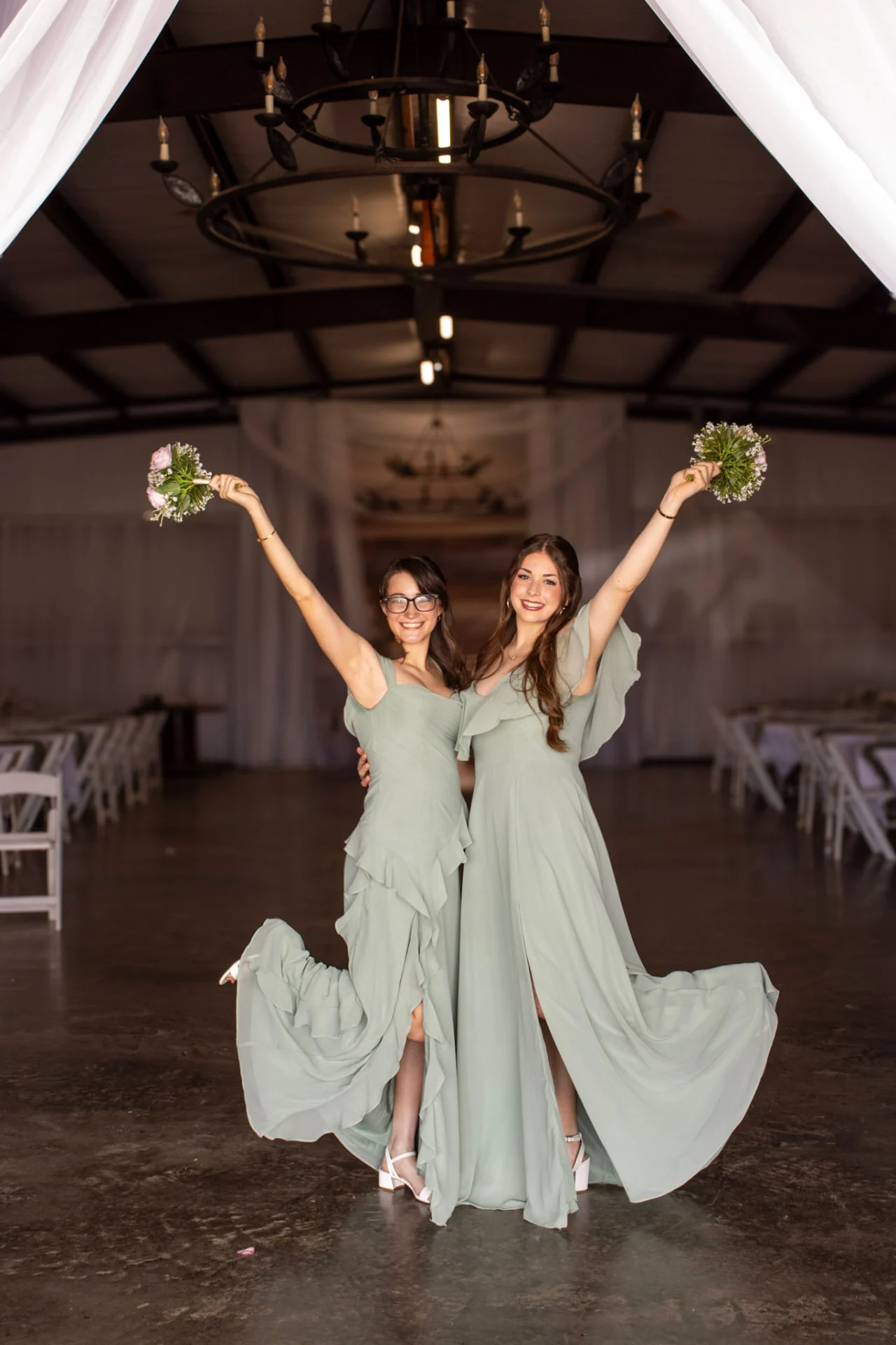Two bridesmaids in sage green gowns raising their bouquets inside the draped reception hall