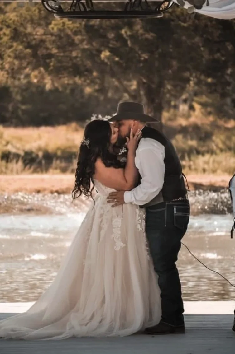 Bride and groom in a cowboy hat sharing their first kiss on the lakeside dock with the fountain behind them in Normangee, Texas