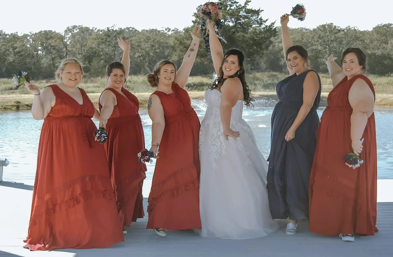 Bride and five bridesmaids in red and navy dresses raising their bouquets on the dock beside the lake