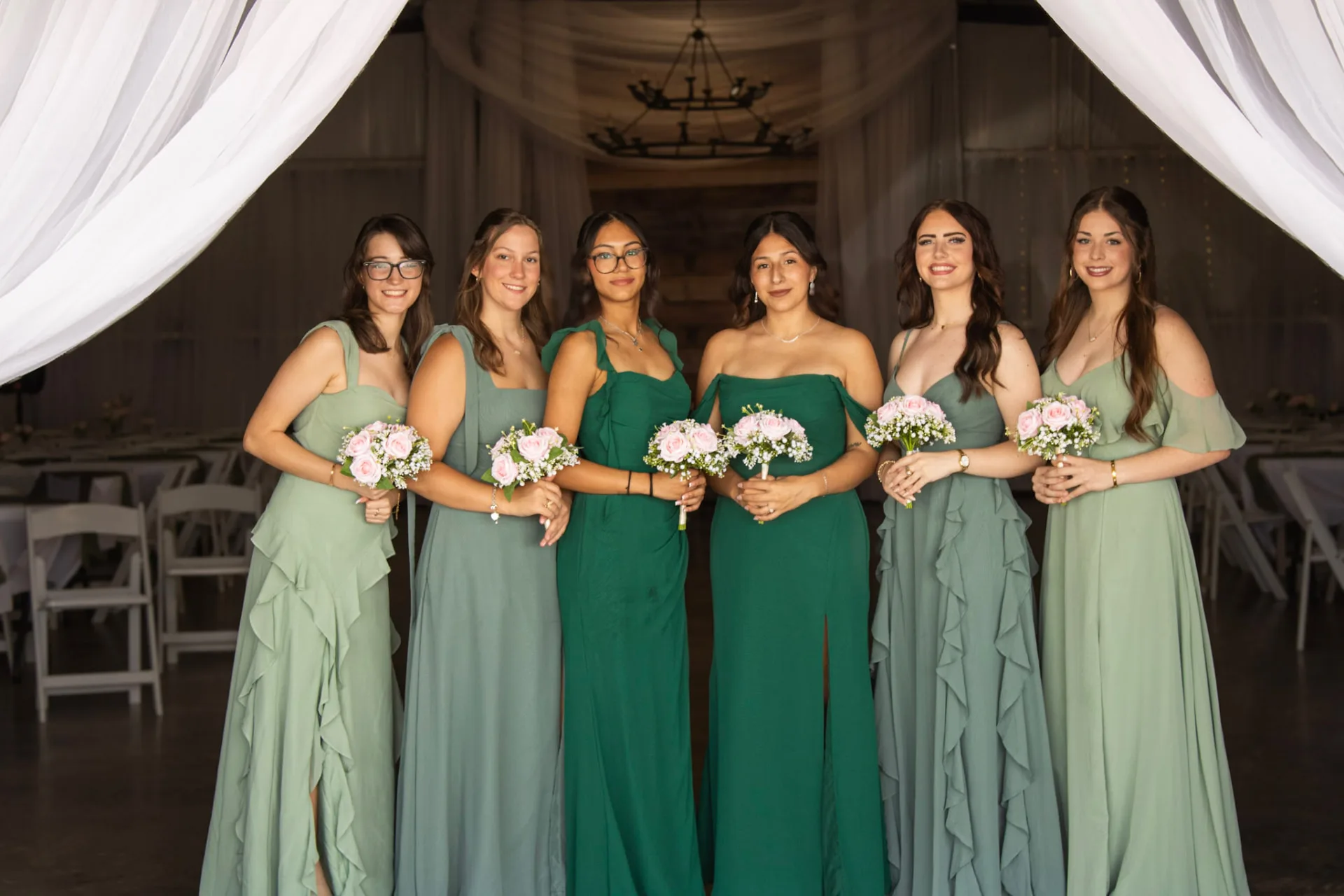 Six bridesmaids in sage and emerald gowns holding blush bouquets in the draped barn entry