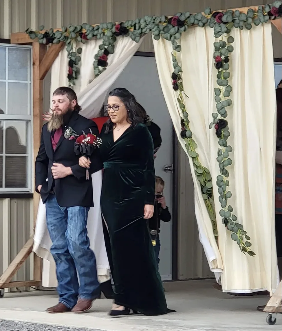 Groomsman in a black suit escorting a guest in an emerald velvet gown under the eucalyptus and rose garland arch at Reverie at Diamond D