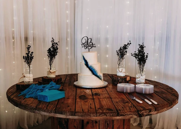 White wedding cake with a teal geode design on a rustic spool table against a twinkle-light backdrop