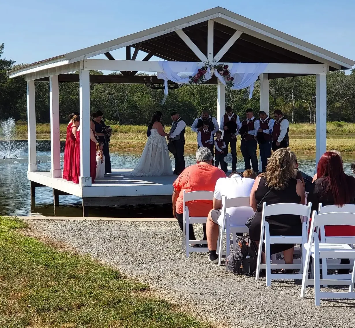 Guests seated in white chairs watching a prayer during a lakeside ceremony with a water fountain behind the pavilion