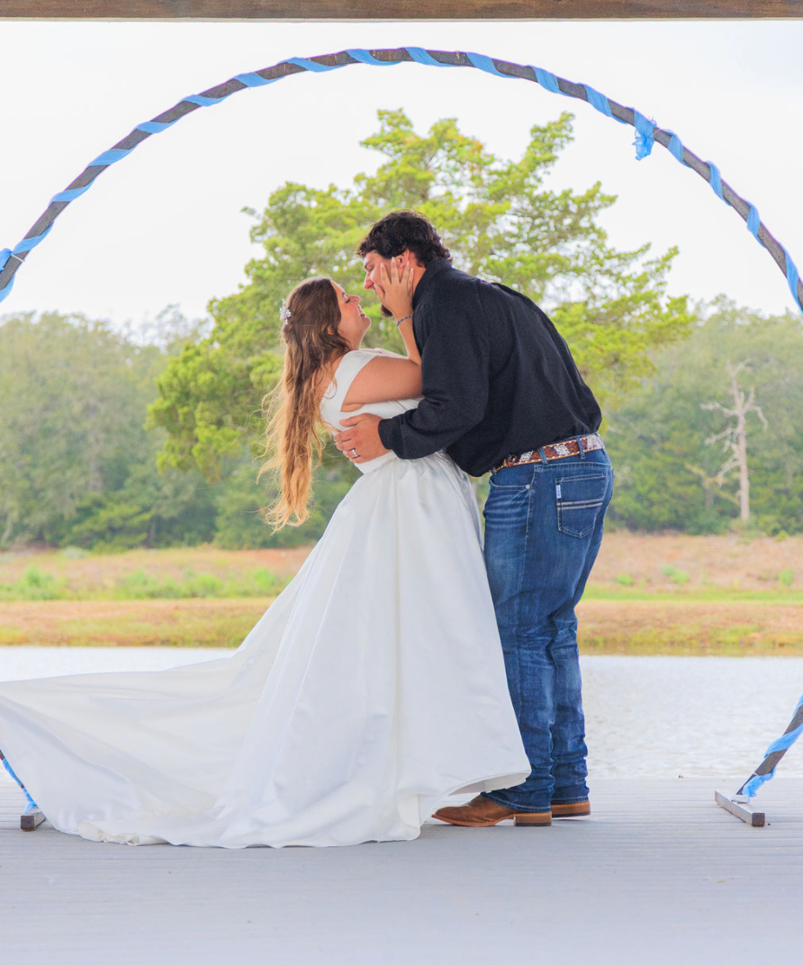 Jaelyn & Keelan's wedding at Reverie at Diamond D — the bride and groom sharing a kiss under the blue-wrapped circular arch over the lake